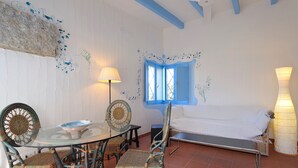 Una habitación con una mesa redonda de vidrio, sillas de mimbre, un sofá blanco, una ventana con contraventanas azules y un ventilador de techo.