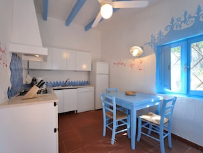 Eine Küche mit weißen Schränken, einem blauen Tisch und Stühlen, einem Kühlschrank und einem Fenster mit blauen Fensterläden.