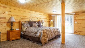 A bedroom with a wooden ceiling and walls, a bed with patterned bedding, a nightstand with a lamp, and a window with blinds.
