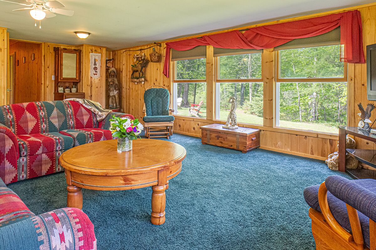 Interior acolhedor de uma cabana, com decoração em madeira, uma mesa de centro redonda também de madeira, uma poltrona azul e um sofá com um cobertor colorido.
