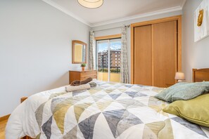 Une chambre avec un grand lit, une armoire en bois, une table de chevet, une lampe, une fenêtre avec des rideaux et une vue sur les bâtiments à l’extérieur.
