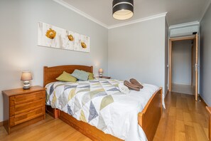 Een slaapkamer met een houten bed, een nachtkastje, een lamp, een decoratief kunstwerk aan de muur en een deur die naar een andere kamer leidt.
