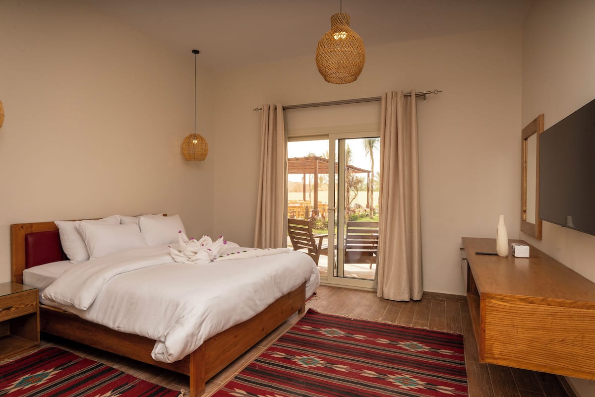 A bedroom with a bed, a wooden desk, a large mirror, and a view of the outdoors.