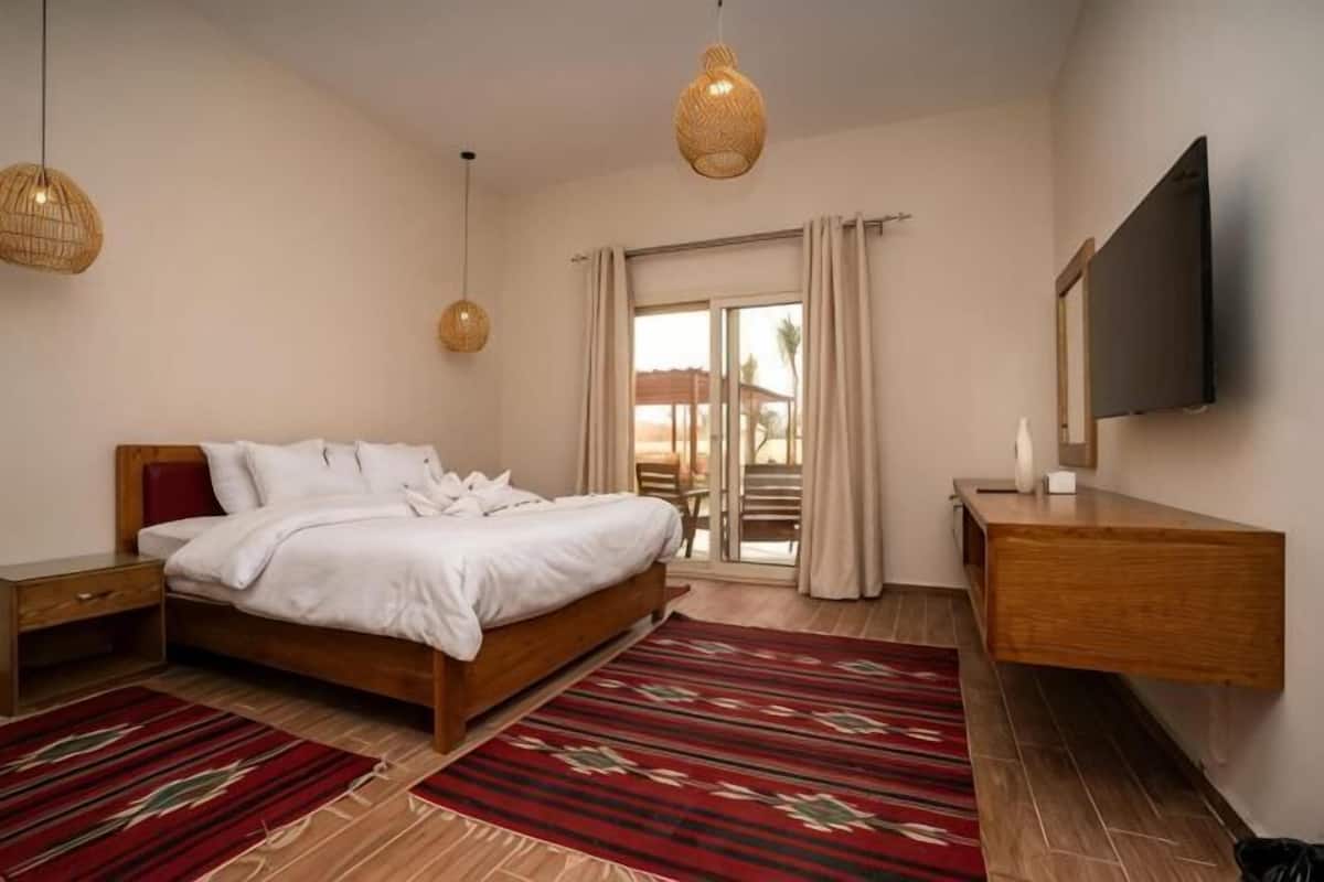 A bedroom with a bed, bedside tables, a TV, a wooden dresser, and a balcony with curtains.