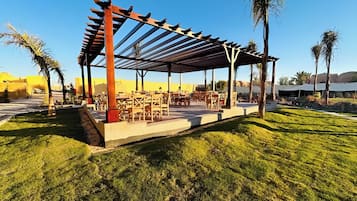 Ulkoterassi, jossa on pöytiä ja tuoleja ja jonka yläpuolella on pergola. Alue on nurmikon ja palmujen ympäröimä.