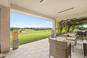 Un patio couvert aménagé avec du mobilier d’extérieur, offrant une vue sur un paysage verdoyant et agrémenté d’un pilier en briques.