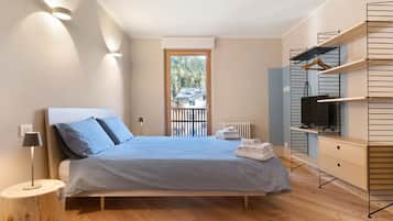 Une chambre moderne avec un lit, une lampe de chevet, un plancher en bois, une étagère intégrée pour la télévision et une vue sur l’extérieur.