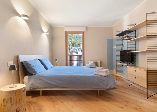 A modern bedroom with a bed, bedside lamp, wooden floor, built-in TV shelf, and a view of the outdoors. - Apartment 'Palazzo Righi - Blu' with Mountain View, Balcony and Wi-Fi (Carisolo)