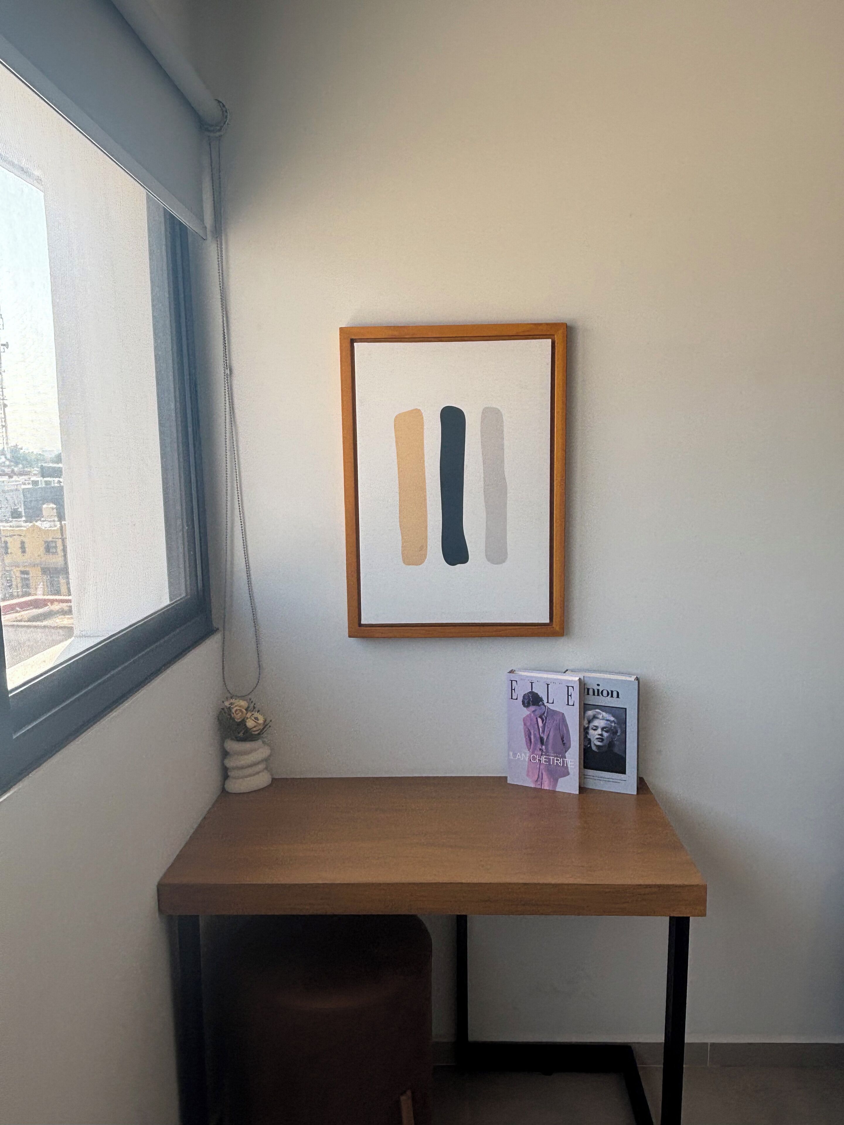 A corner room with a wooden desk, a framed abstract artwork, and a vase with dried flowers.
