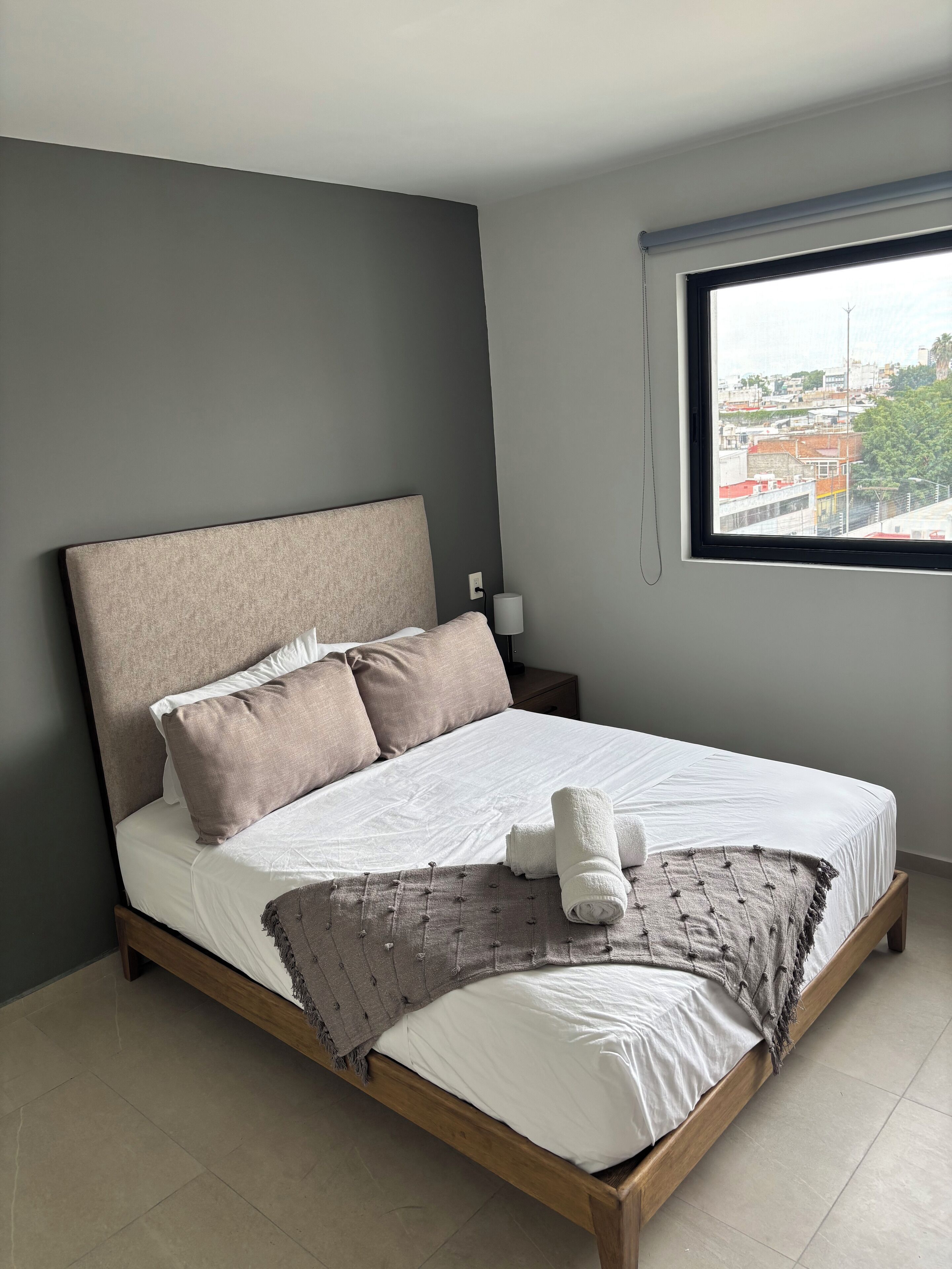 A neatly made bed with a wooden frame, a grey headboard, and a view of a cityscape through a window.