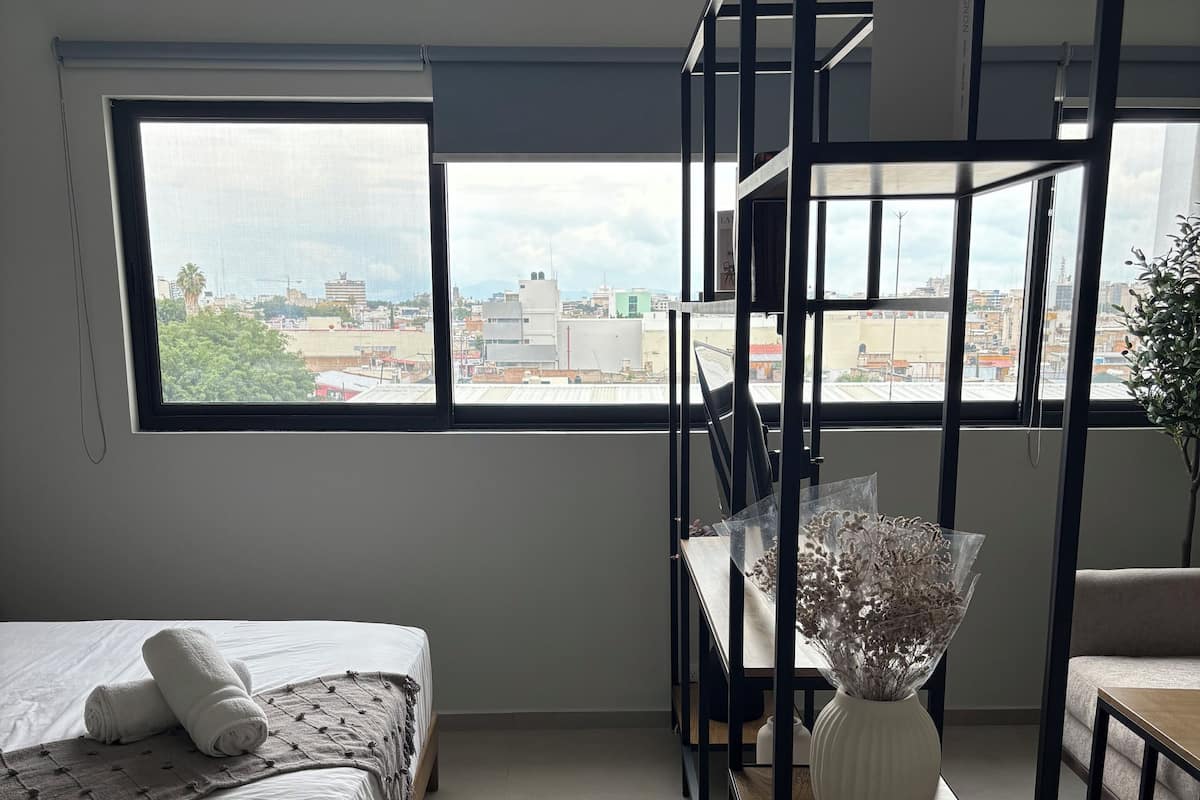 A modern bedroom with a bed, a desk, and a large window offering a city view.