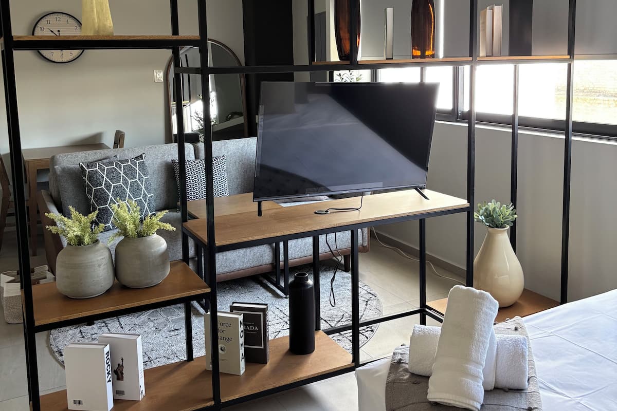 A modern living room with a TV on a wooden stand, a ceiling fan, and a large window providing natural light.