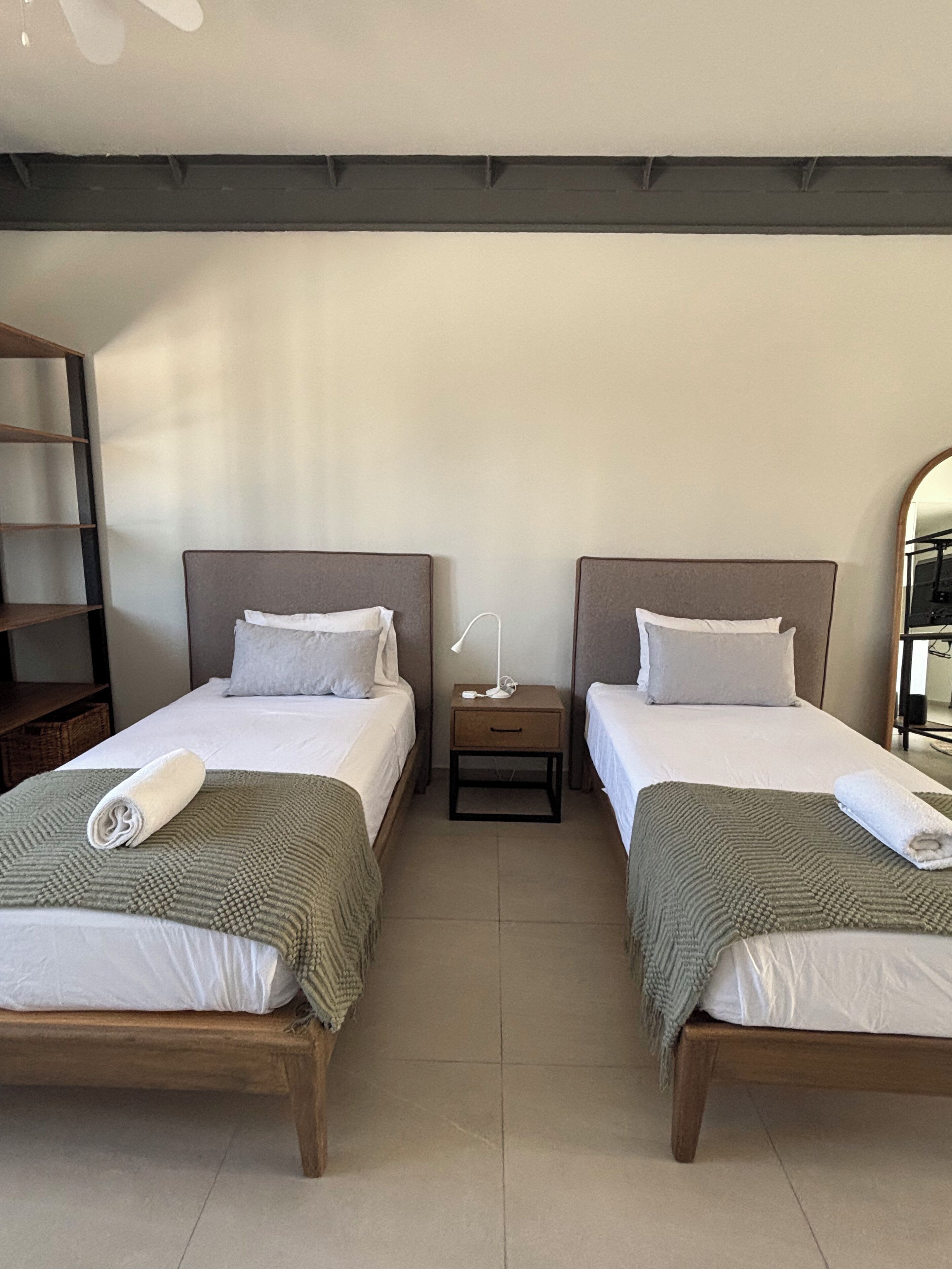 Habitación de hotel con dos camas individuales, cada una con una almohada y una toalla enrollada. Hay una mesita de noche entre las camas con una lámpara y un cajón pequeño. El piso es de madera y hay una estantería a la izquierda.
