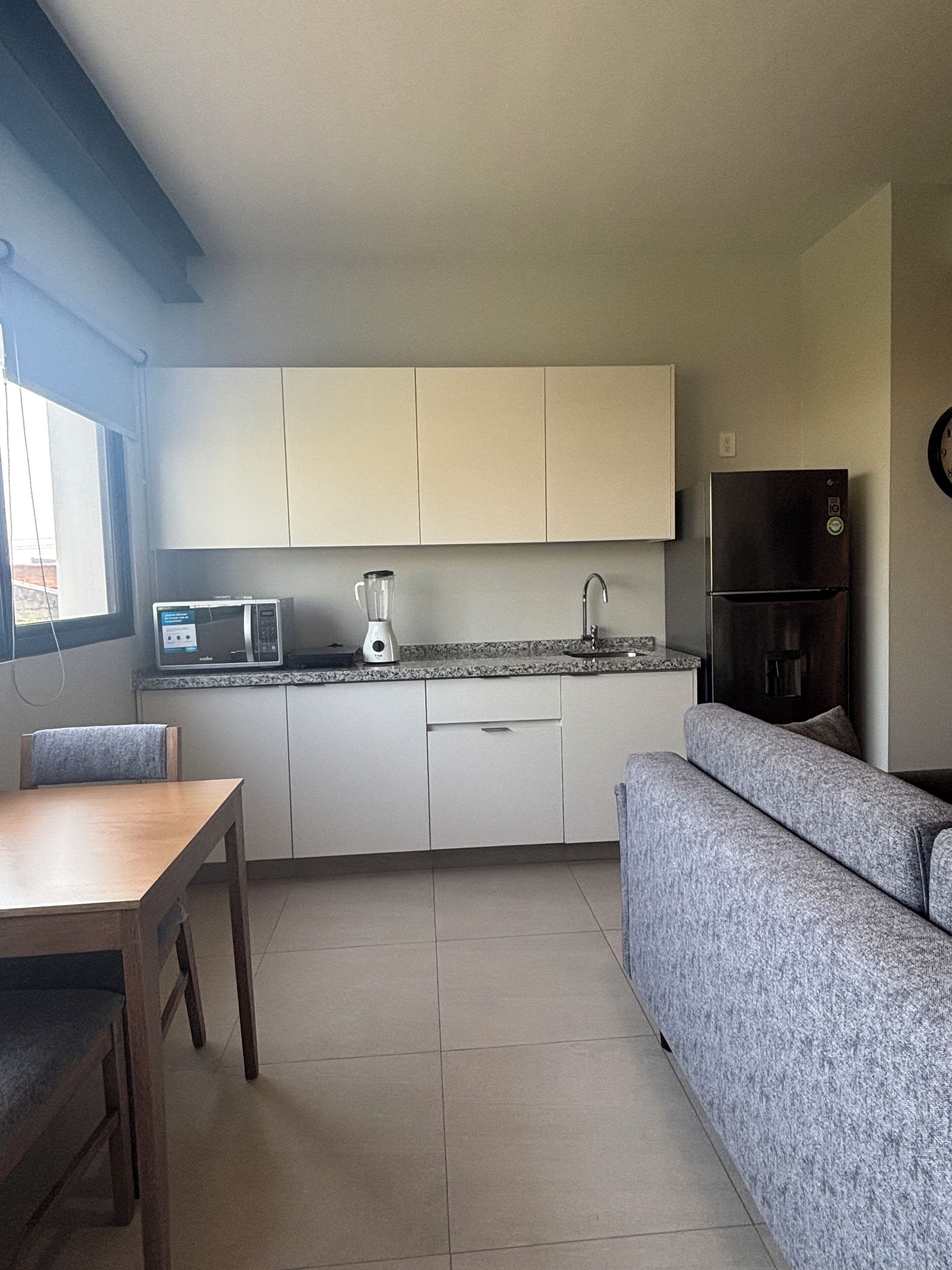 Una cocina compacta con armarios blancos, microondas, licuadora, refrigerador y un sofá gris.