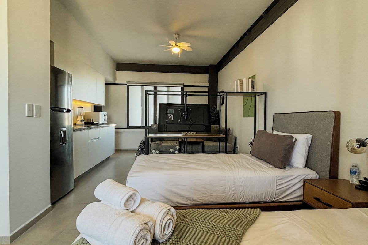 A compact hotel room with a kitchenette, a bed with a headboard, and a green textured rug.