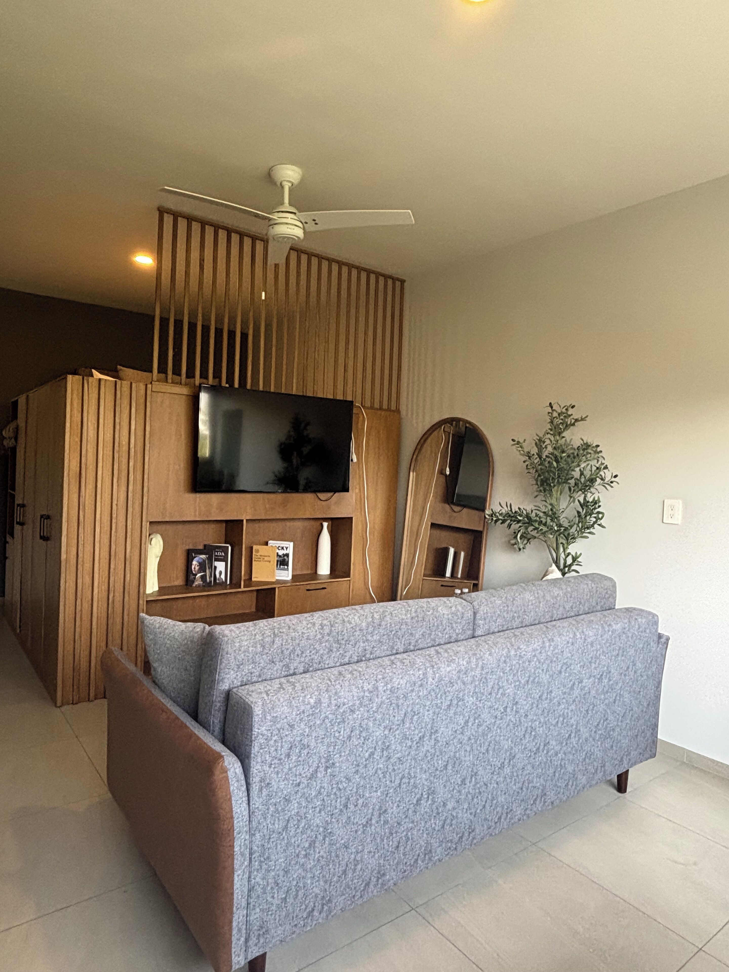 A modern living room with a grey sofa, wooden paneling, a ceiling fan, a flat-screen TV, and a potted plant.