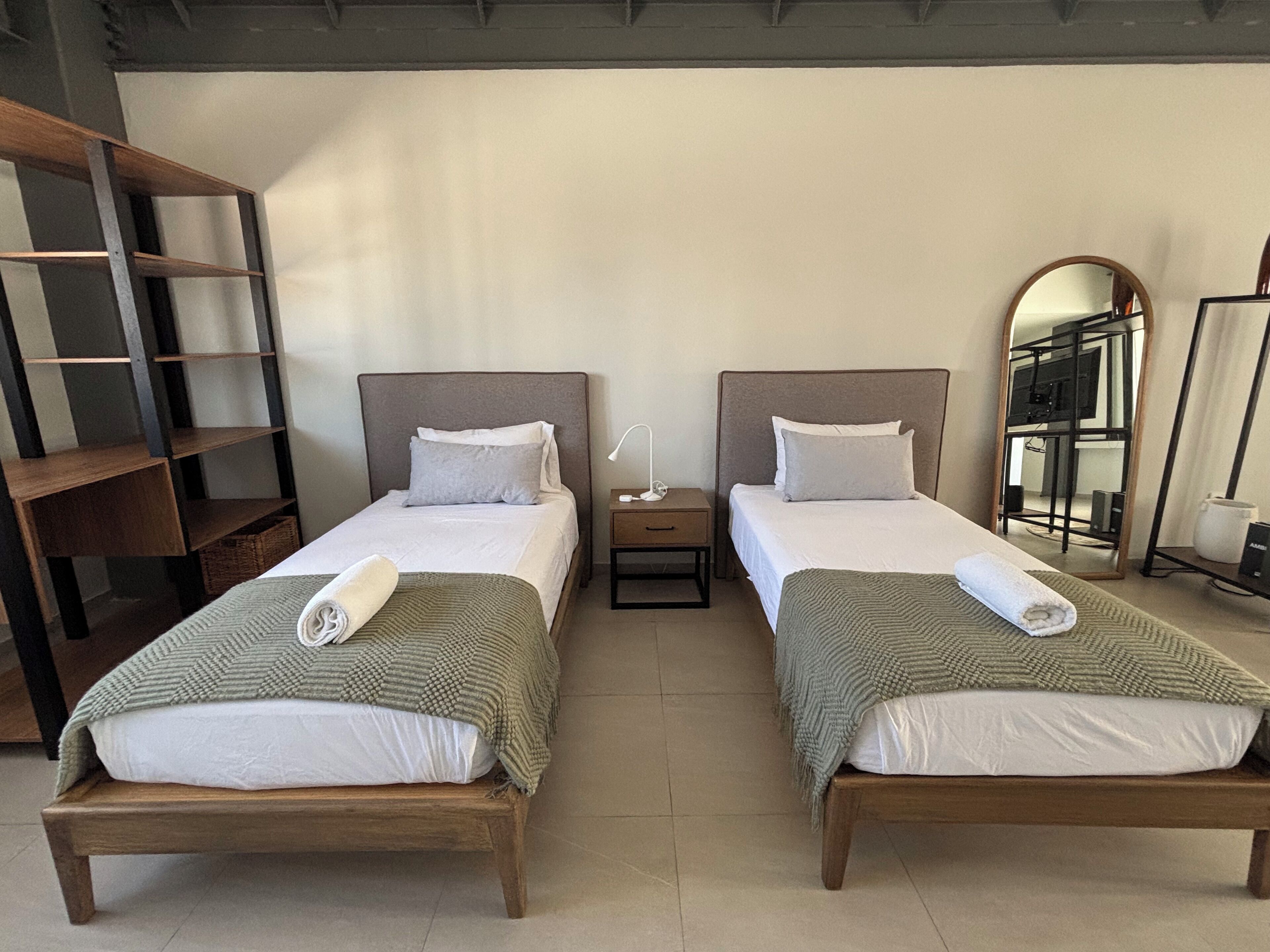 Habitación de hotel con dos camas individuales, cada una con estructura de madera, ropa de cama blanca y una manta verde. Se observa un mesita de noche con lámpara, un espejo y una estantería a la izquierda.