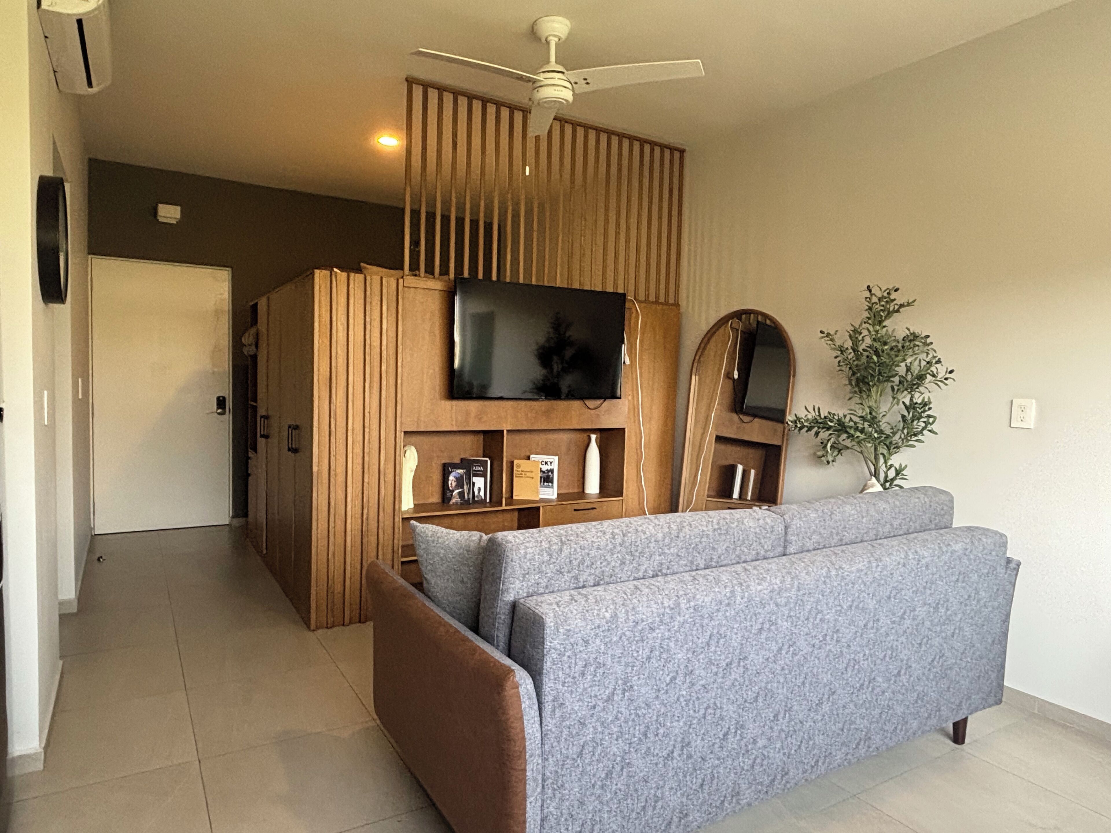 A modern living room with a grey sofa, wooden paneling, a ceiling fan, a flat-screen TV, and a potted plant.