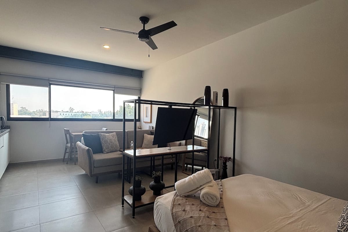 A modern bedroom with a bed, a desk, and a chair. There is a window providing natural light and a view of the outside.