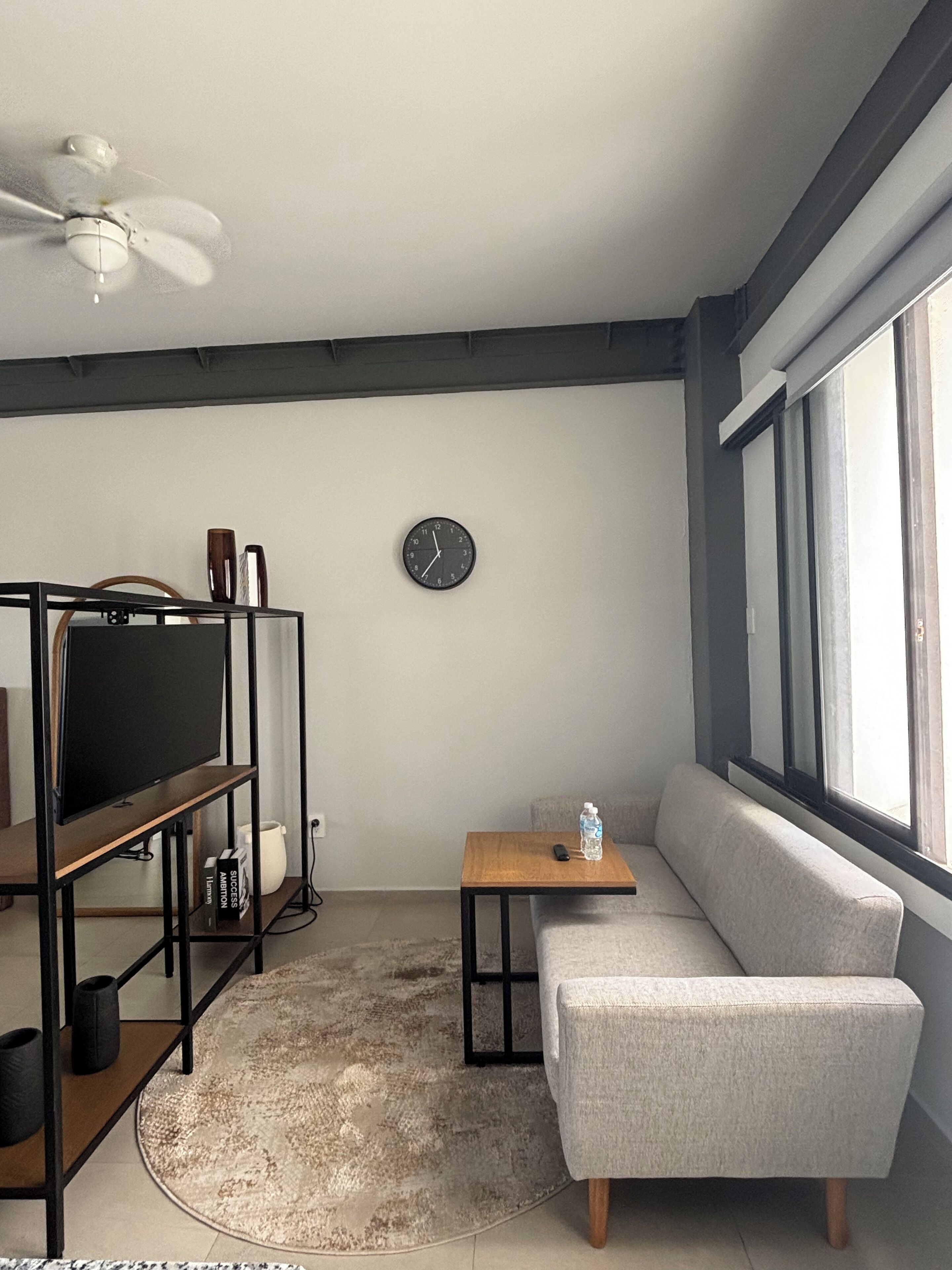 Un salon moderne avec un téléviseur posé sur un meuble, un canapé, une petite table avec une bouteille, une horloge au mur et un ventilateur de plafond.