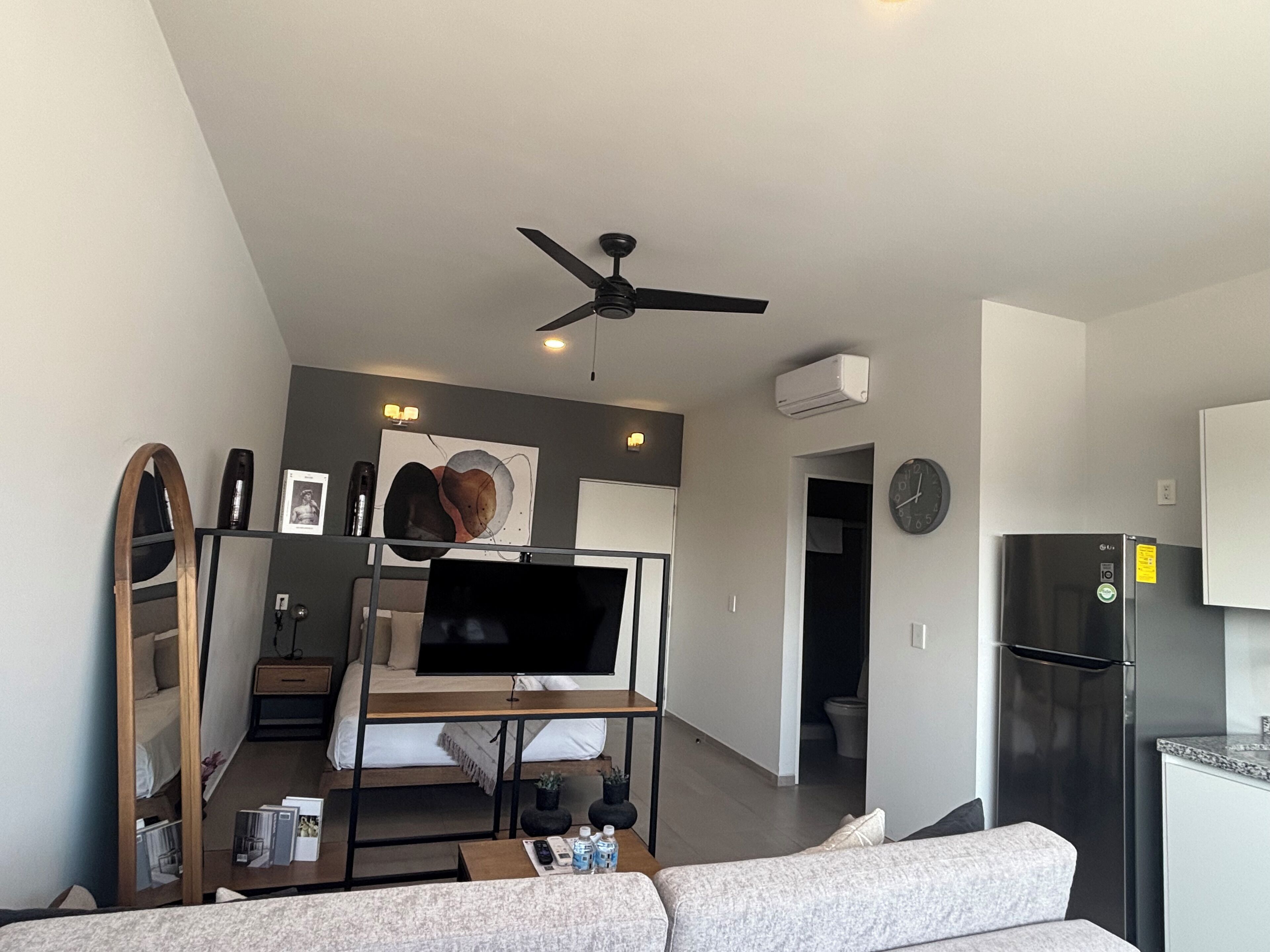 A modern living room with a flat-screen TV, a ceiling fan, and a kitchen area with a refrigerator.