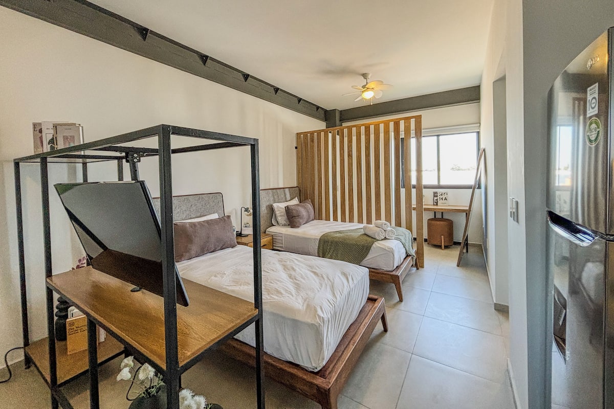 A modern bedroom with a four-poster bed, two single beds, a wooden partition, a ceiling fan, a refrigerator, and a window with blinds.