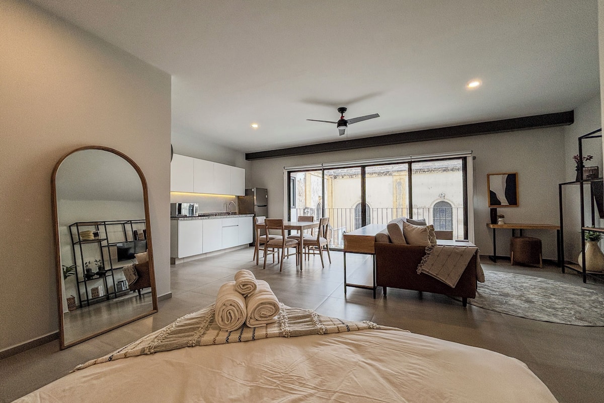 A modern bedroom with a large bed, a dining area, and a kitchen in the background.