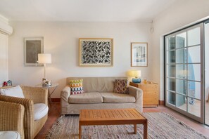 A living room with a sofa, a coffee table, a side table with a lamp, and a large window with a view. - Villa 'Casa Do Ouriço - Cabanas' with Sea View, Wi-Fi and Air Conditioning (Tavira)