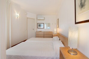 A hotel room with a double bed, a bedside table with a lamp, a dresser, a mirror, and an air conditioning unit on the wall.