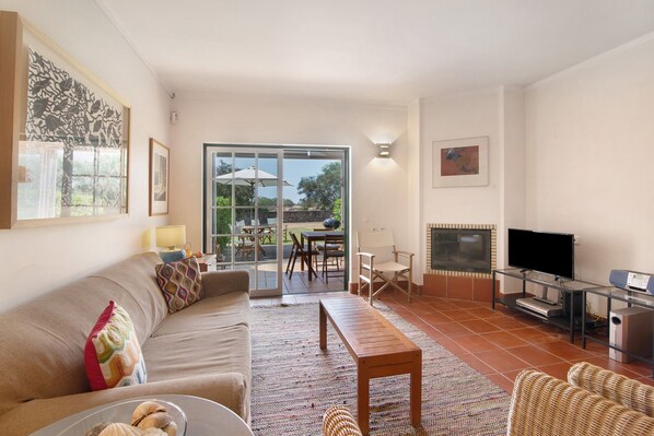 A living room with a sofa, a coffee table, a TV, and a fireplace. There is a sliding glass door leading to an outdoor patio with a table and chairs.
