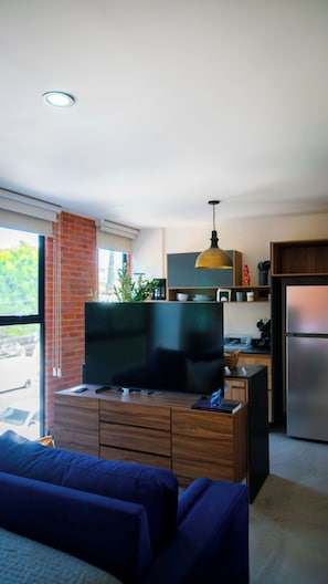 A modern living room with a flat-screen TV, a sofa, and a dining area with a refrigerator and wooden cabinets. - Americana apartment with rooftop pool (Guadalajara)