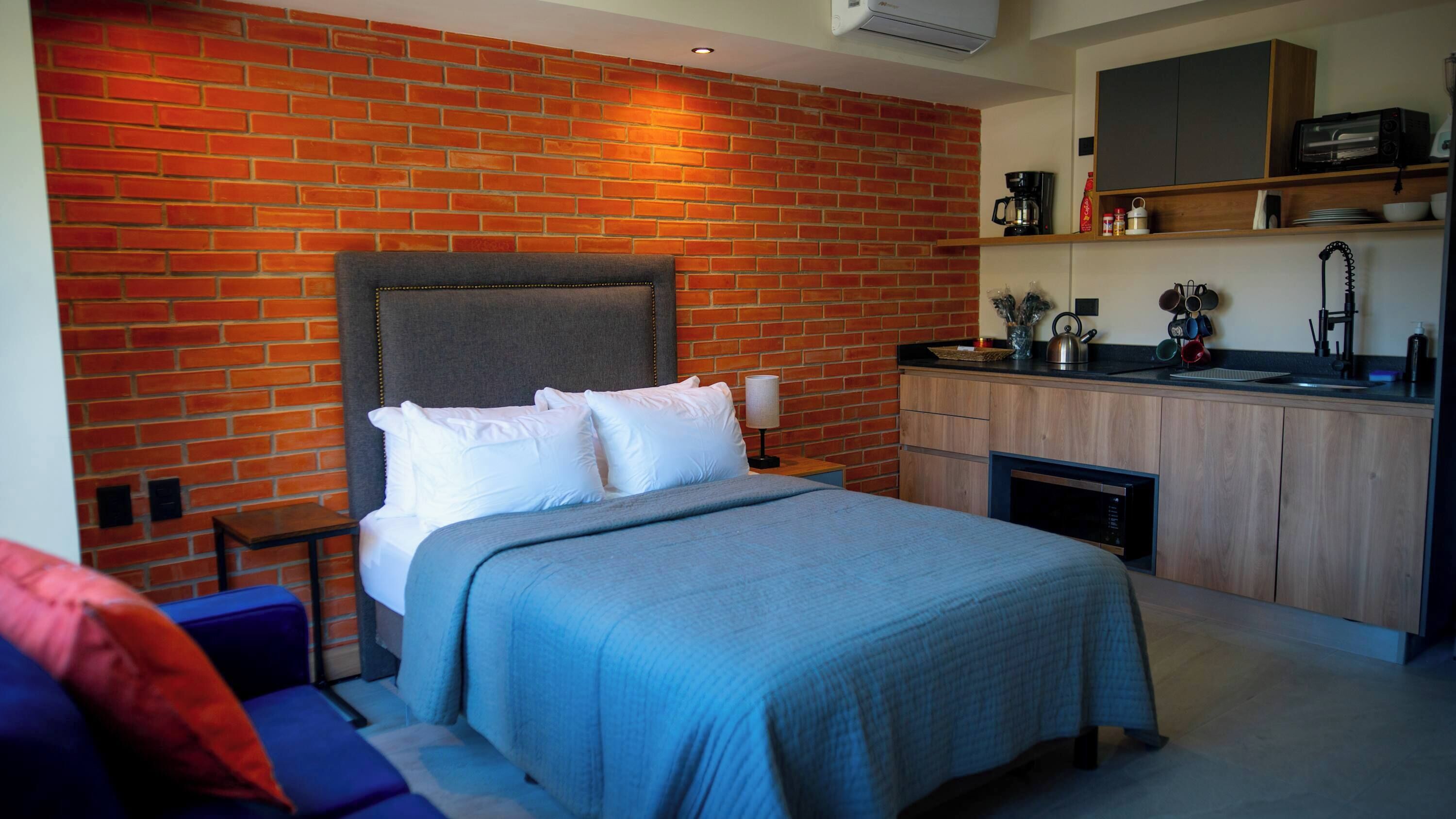 Quarto com parede de tijolos, cama com roupa de cama branca, cabeceira cinza e uma pequena cozinha compacta com micro-ondas e cafeteira.