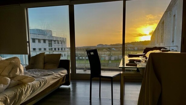A room with a bed, a chair, and a table. There is a large window providing a view of buildings and a sunset. - São Pedro Apartment (Vila Franca Do Campo)