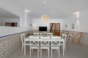 A dining area with a white table and chairs, a hanging light fixture, a television, and a tiled wall with a blue border.