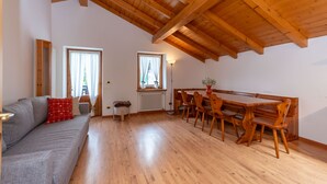 A spacious room with a wooden ceiling, a grey sofa with red cushions, a dining table with wooden chairs, and two windows with curtains. - La Casa di Elvira-Mansarda (Alleghe)