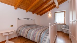 A bedroom with a wooden ceiling, a bed with striped bedding, a bedside table, a window with curtains, and a radiator. - La Casa di Elvira-Mansarda (Alleghe)