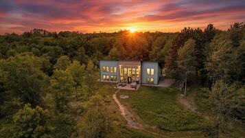 A modern two-story house surrounded by dense forest, with a clear path leading to the entrance.