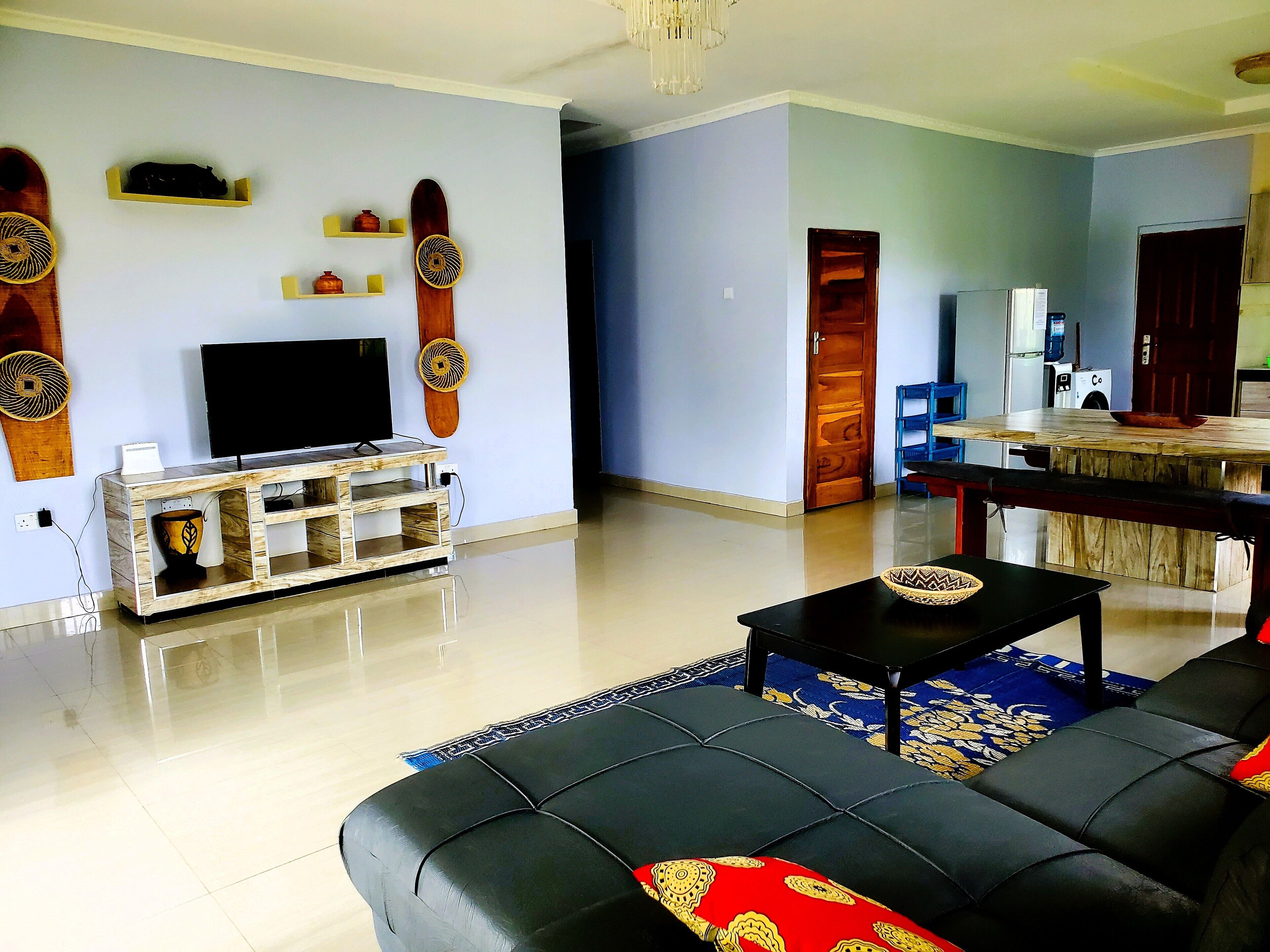 Sala de estar con un sofá negro, un televisor sobre un mueble de madera, una zona de comedor con mesa de madera, y una cocina visible a través de una puerta abierta.