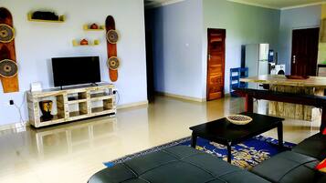 Sala de estar con un sofá negro, un televisor sobre un mueble de madera, una zona de comedor con mesa de madera, y una cocina visible a través de una puerta abierta.