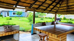 An outdoor covered seating area with a wooden table and benches, a thatched roof, and a view of a blue house and garden. - Stylish Home w/Private Pool, AC and Free Starlink High-speed Wi-Fi (Lusaka)