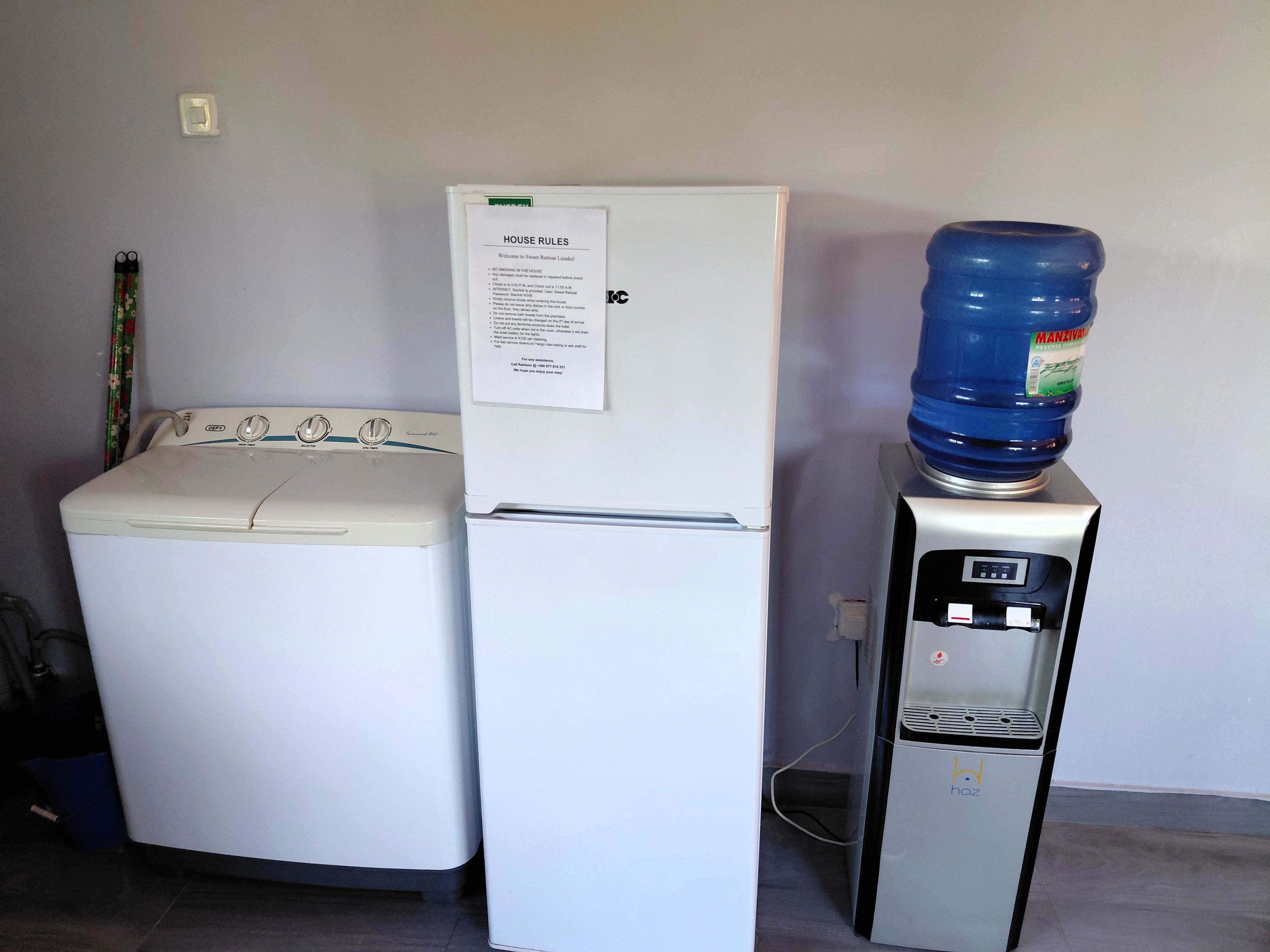 Une machine à laver blanche, un réfrigérateur blanc sur lequel est affiché un panneau indiquant les règles de la maison, et un distributeur d’eau.