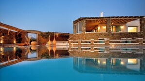 A modern poolside area with stone walls, a wooden deck, and a clear pool reflecting the surroundings. - Villa Irida (Mikonos)