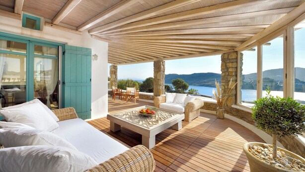 A sunlit outdoor living area with a wooden deck, wicker furniture, and a stone pillar. - Villa Irida (Mikonos)