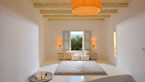 A bedroom with a large bed, a window with shutters, bedside lamps, and a hanging light fixture. - Villa Irida (Mikonos)
