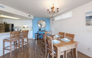 A dining area with a wooden table set for four, surrounded by wooden chairs. A chandelier hangs above the table, and a kitchen with white cabinets is visible in the background. - Seascape Dream! Bella Vista North Bluff Oasis (Aptos)