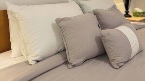 Ein ordentlich bezogenes Bett mit weißen und grauen Kissen, zwei gerahmte Wandbilder, eine Nachttischlampe und ein Fenster mit Jalousien.