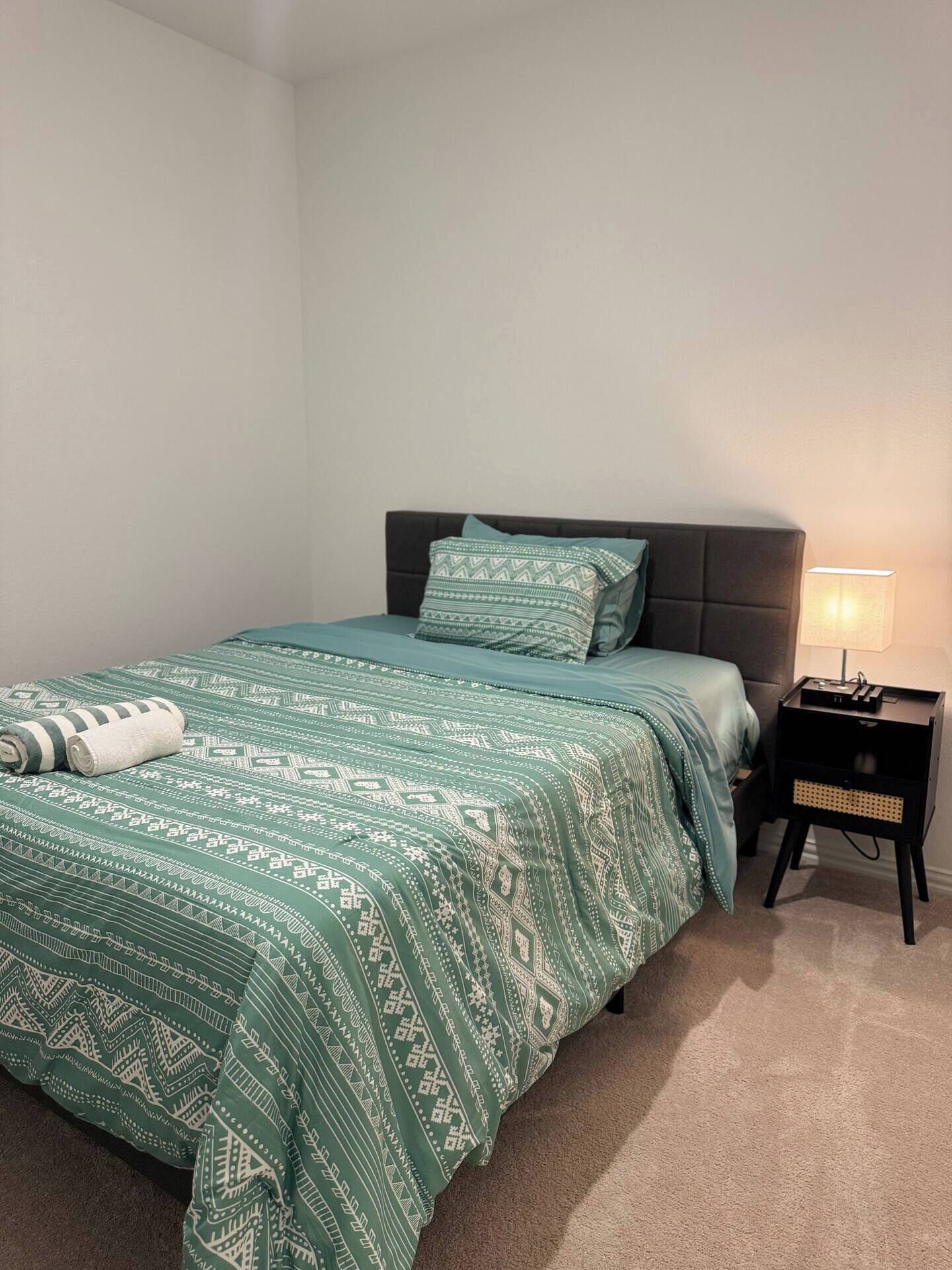 Ein ordentlich bezogenes Bett mit einer gemusterten grünen Tagesdecke und Kissen, ein kleiner Nachttisch mit Lampe und eine schlichte Wand.