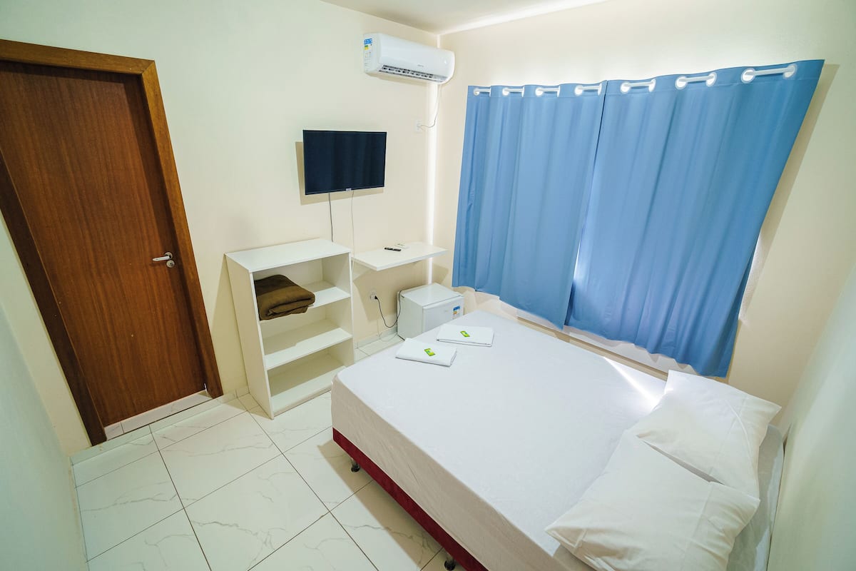 A hotel room with a bed, a TV, a shelf, and a window with curtains.