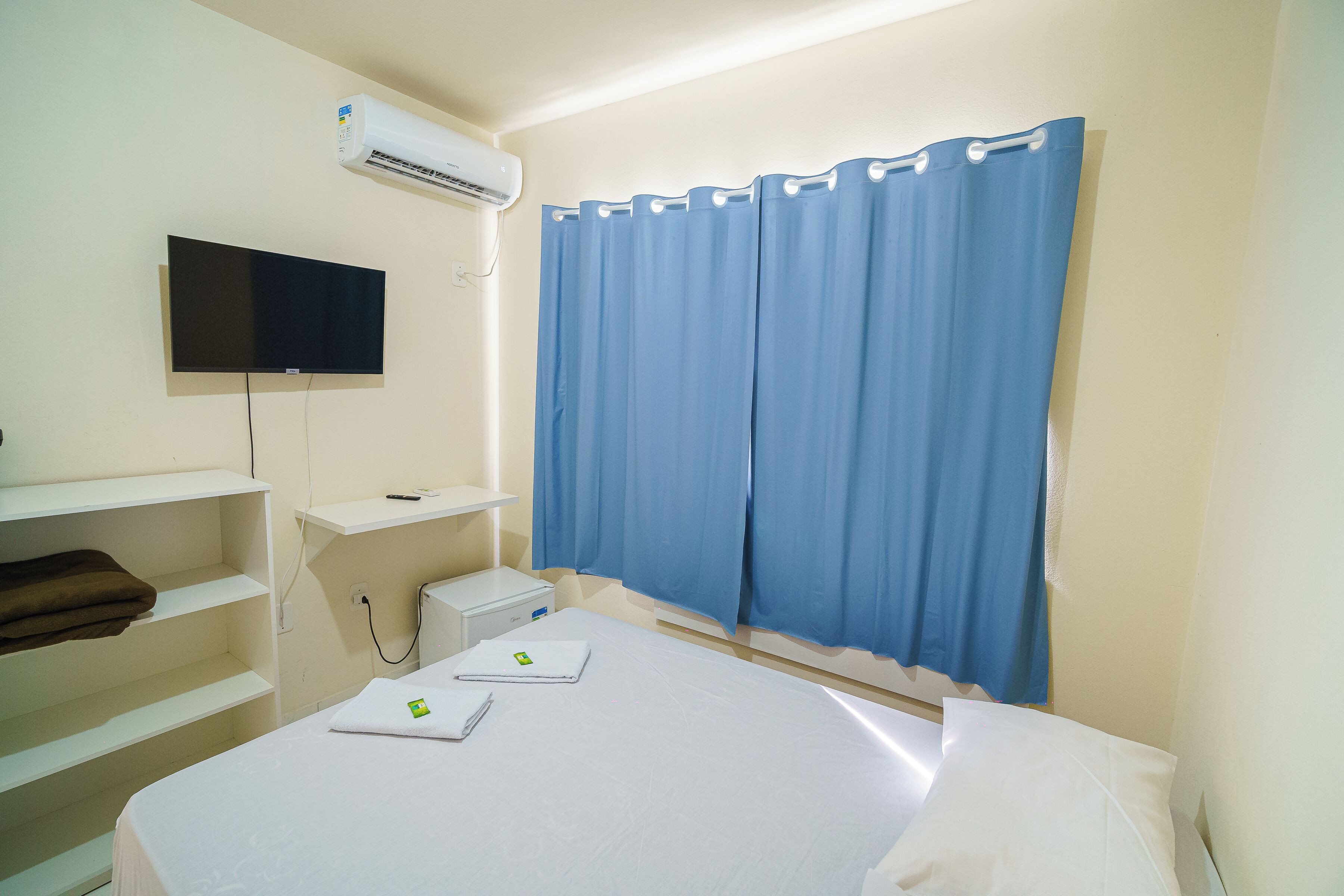 Ein Hotelzimmer mit Bett, flachbildfernseher an der Wand, Schreibtisch mit Computer und blauem Vorhang.