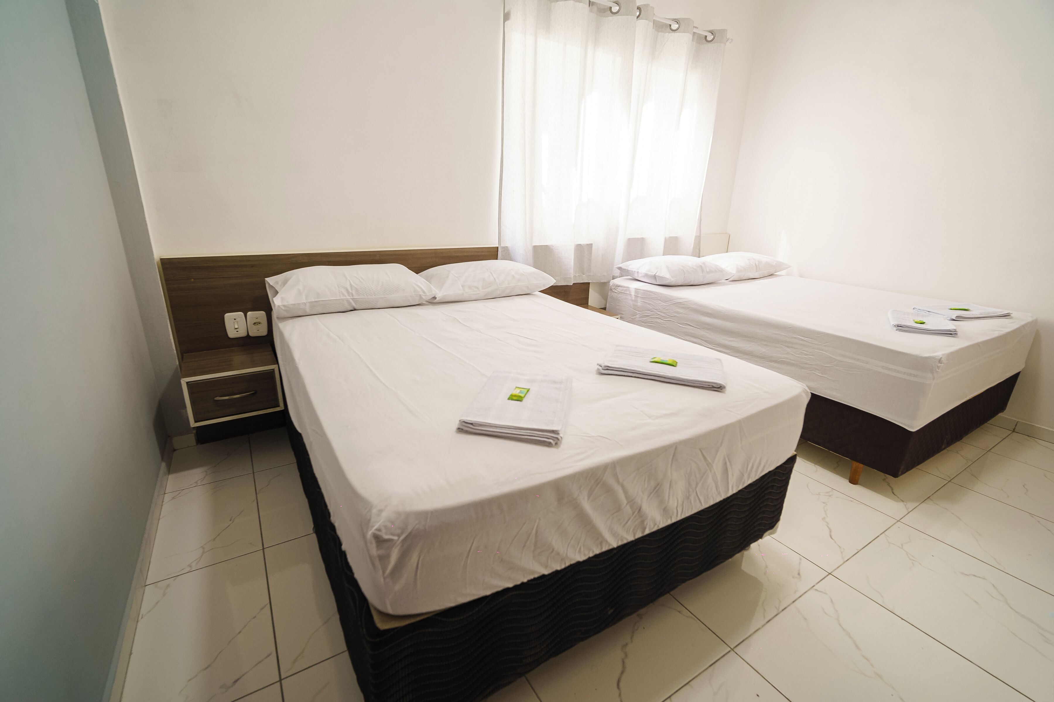 A hotel room with two single beds, a nightstand, a small drawer, and a window with sheer curtains.